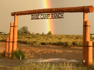 A wooden entrance sign for Base Camp Ranch stands in front of a field with a rainbow in the background.