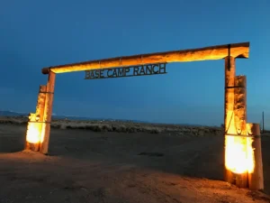 Large wooden entrance sign reading "Base Camp Ranch" lit by yellow lights at dusk in a desert landscape.