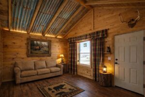 Rustic cabin room with wood walls, a plaid-curtained window, sofa, nature decor, and antlers mounted on the wall.