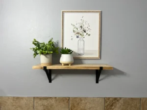 A wooden shelf with two potted plants is mounted on a gray wall beneath a framed floral artwork.