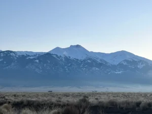 Snow-capped mountains rise above a dry grassy plain under a clear sky with soft morning or evening light.