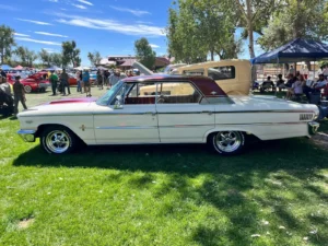 A white and maroon classic car is parked on grass at an outdoor car show with people and other vehicles nearby.