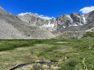 Rocky mountains with patches of snow rise above a grassy alpine meadow and small pools under a clear blue sky.