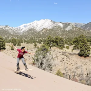Person sandboarding down a sand dune with mountains and trees in the background under a clear blue sky.