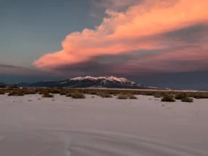 Snow-covered field with sparse shrubs, distant mountains, and a large pink cloud against a darkening sky at sunset.