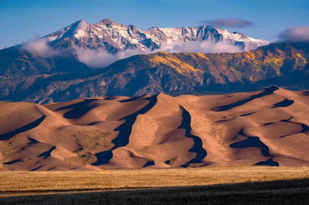 Sand dunes in the foreground with forested hills and snow-capped mountains under a blue sky with scattered clouds.
