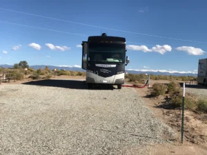 A motorhome parked on a gravel lot at a campsite with mountains and clear skies in the background.
