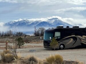 A motorhome is parked in a gravel lot with snow-capped mountains and cloudy skies in the background.